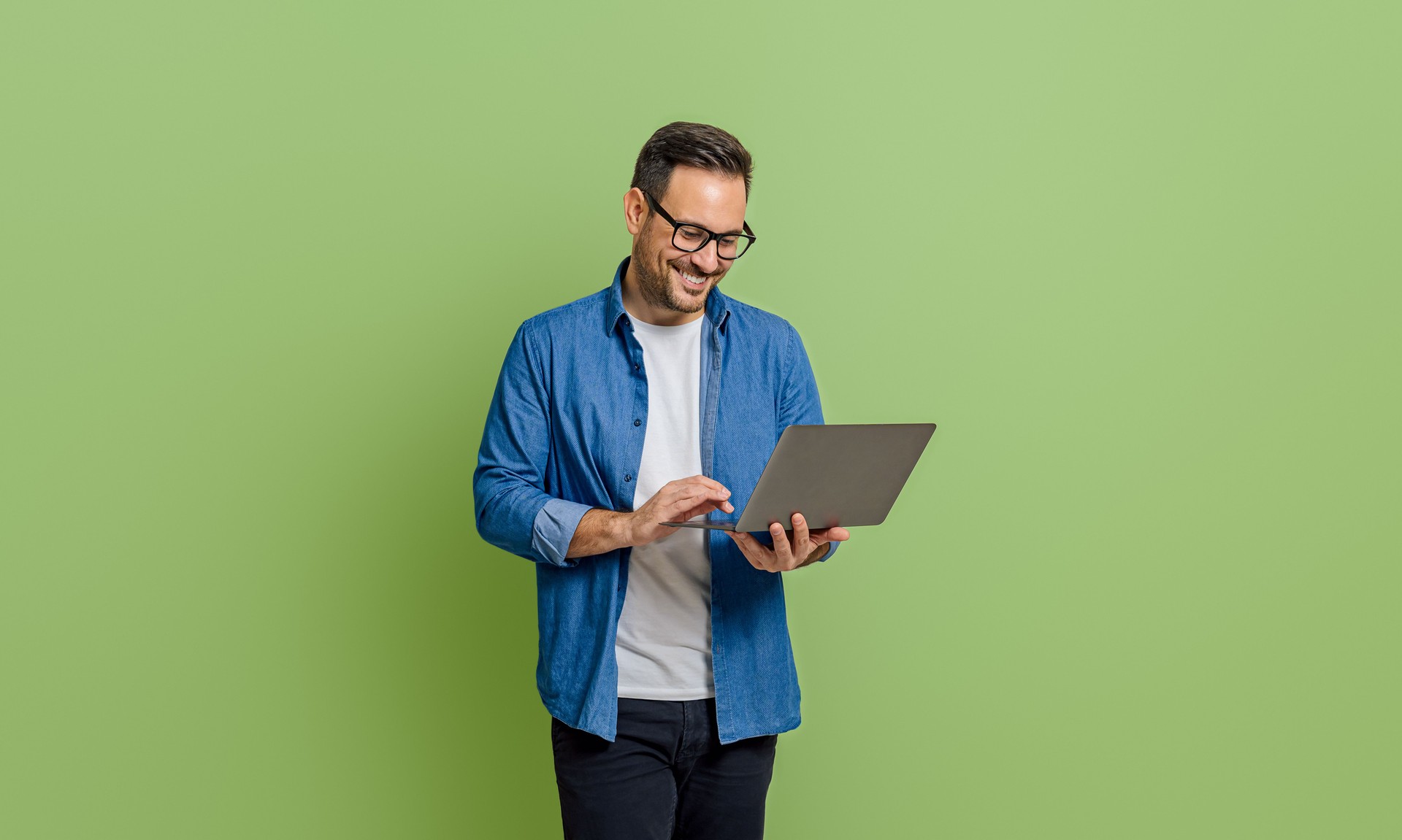 Smiling young businessman analyzing data over laptop while standing isolated on green background